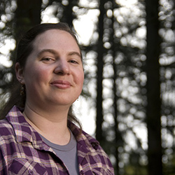 Kathryn Ringland is a white woman with dark brown hair pulled off her face, smiling at the camera. She is wearing a purple shirt with a purple plaid shirt on top.