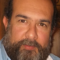 Headshot of Dr Alexandros Pino, a Native American man living in Greece. He wears a light blue shirt, and he has dark hair and a long beard.