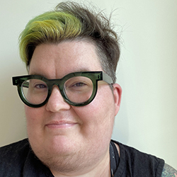 Katta Spiel, a white non-binary person with green hair and big glass frames looking amused into the camera with their arms crossed. 