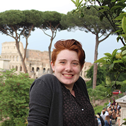 Emma McDonnell, a white woman with short red hair wearing a grey top, smiles at the camera with the Colosseum and foliage out of focus in the the background